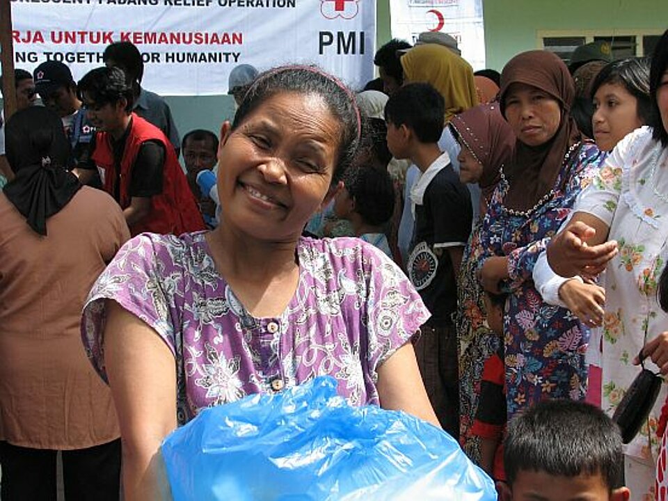 Foto: Lachende ältere Frau mit einem blauen Platsikpaket im Arm, im Hintergrund weitere Personen, die sich am Stand des Indonesischen Roten Kreuzes für Hilfsgüter anstellen