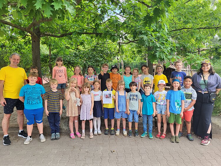 Auf dem Bild stehen viele Schülerinnen und Schüler, sowie Lehr- und Erziehungspersonal der Grundschule am Karpfenteich aus Berlin unter Bäumen und lächeln in die Kamera.