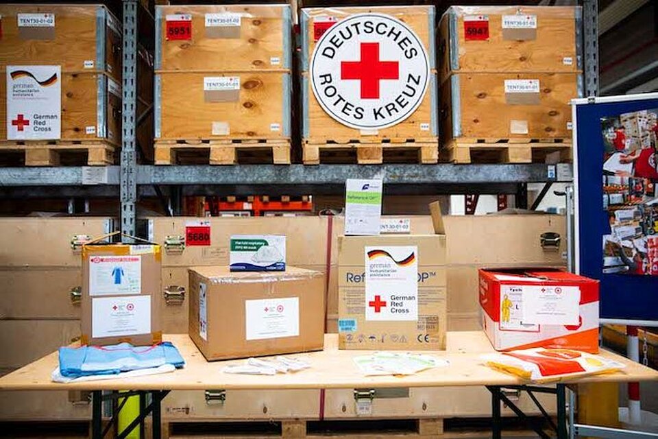 Verschiedene Pakete mit Hilfsgütern stehen vor einem Lagerregal mit weiteren Packkisten und verschiedenen Logos des DRK.
