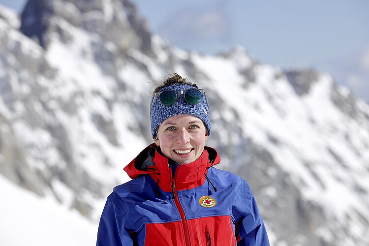 Eine Frau mit einer Mütze und einer rotblauen Jacke mit dem Logo der DRK-Bergwacht vor einer verschneiten Kulisse