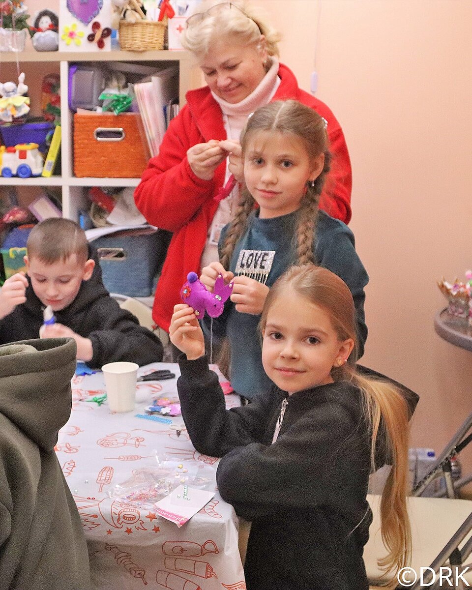 Dieses Bild zeigt drei Kinder und eine erwachsene Frau. Sie alle sitzen und stehen an einem Tisch und basteln im Rahmen einer psychosozialen Fürsorge des Roten Kreuzes in der Ukraine. Die drei Kinder sind etwa 7-10 Jahre alt, zwei Mädchen und ein Junge. Die beiden Mädchen haben langes, glattes dunkelblondes Haar und schauen in die Kamera, der Junge hat kurzes blondes Haar und konzentriert sich aufs Basteln. Die Frau im Hintergrund hat weiße Haare und trägt eine rote Jacke, sie betrachtet interessiert eine Bastelei. Alle vier haben einen fröhlichen Gesichtsausdruck.