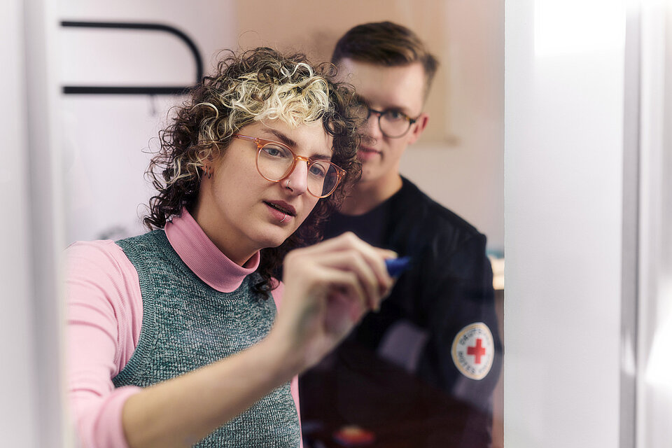Mitarbeiterin des DRK schreibt mit einem Filzstift auf Glas, daneben steht ein Mitarbeiter mit DRK-Abzeichen
