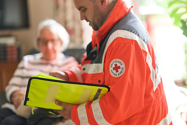 DRKler mit Dokumentenmappe, im Hintergrund Seniorin