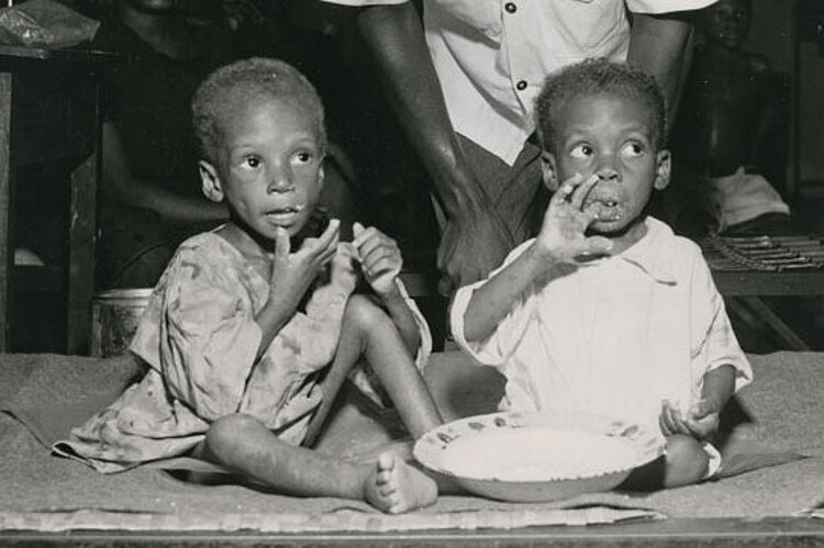 Foto: Ein nigerianischer Rotkreuzhelfer beugt sich über zwei essende Kleinkinder