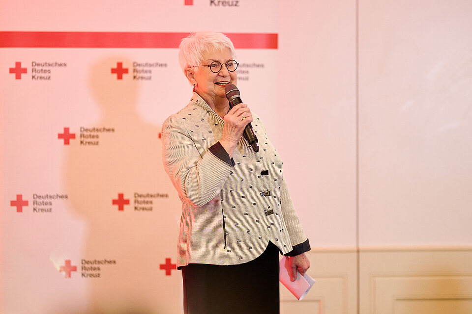DRK-Präsidentin Gerda Hasselfeldt hält ein Mikrofon und spricht vor einem Hintergrund mit dem Logo des Deutschen Roten Kreuzes (DRK). 