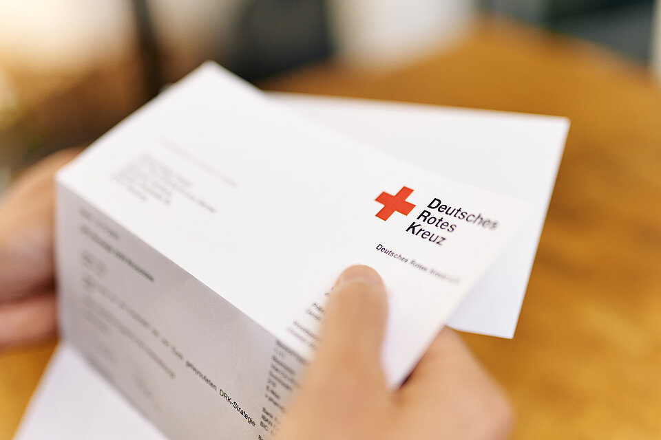 Ein Brief mit DRK-Logo in der rechten Hand gehalten.