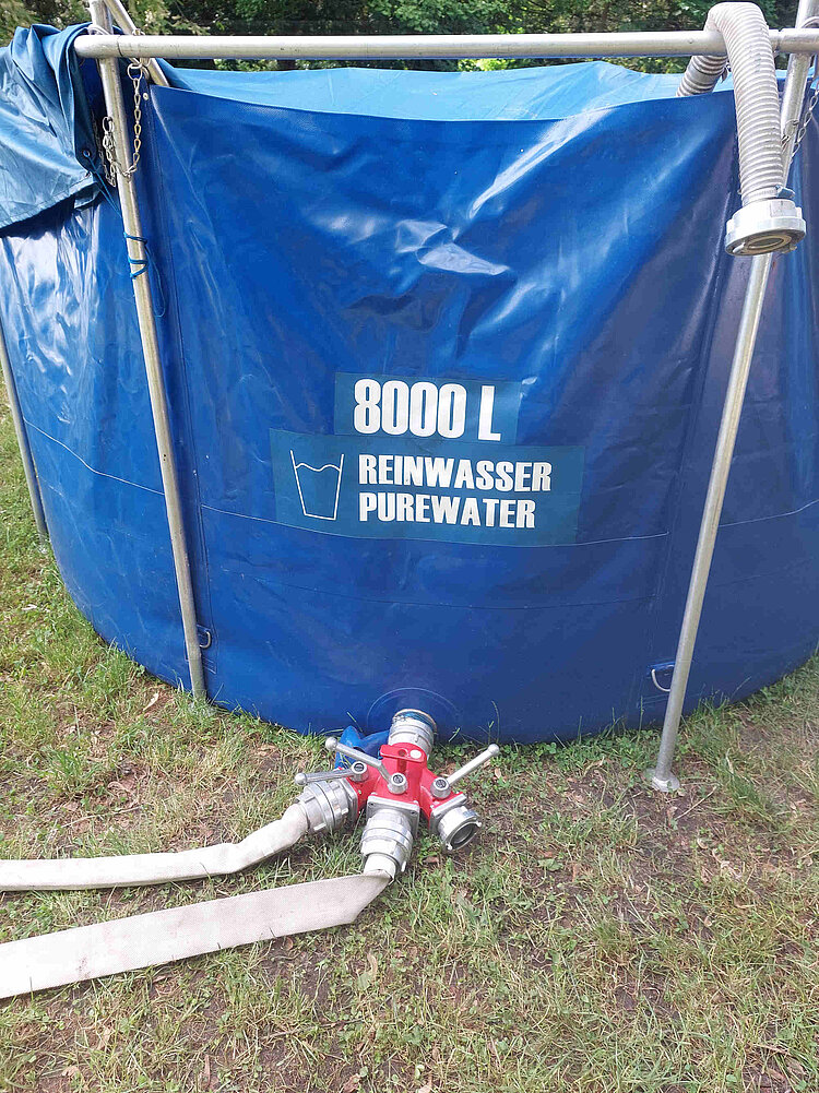 Aus einem Kunststofftank mit der Aufschrift "(8000 Liter Reinwasser Pure Water" ragen mehrere dicke Schläuche