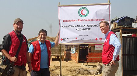 DRK-Mitarbeiter stehen im Flüchtlingslager an einem Banner des Bangladesh Red Crescent