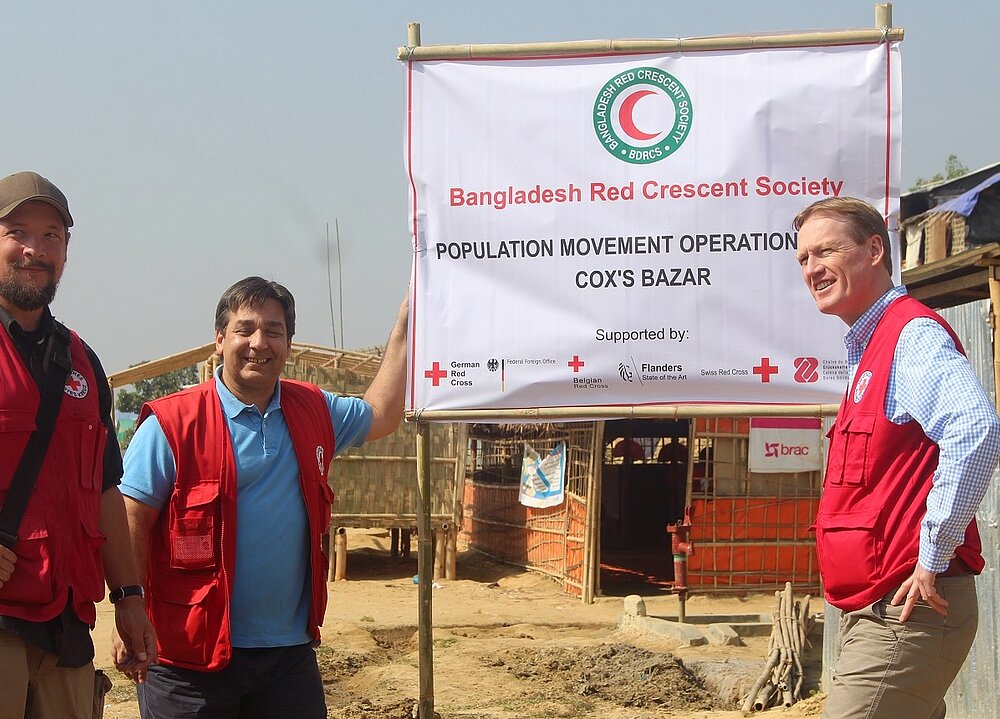DRK-Mitarbeiter stehen im Flüchtlingslager an einem Banner des Bangladesh Red Crescent