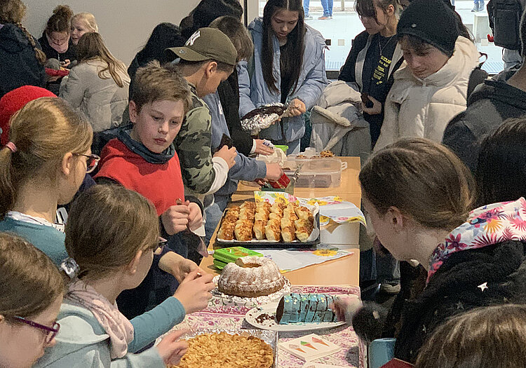 Das Bild zeigt viele Schülerinnen und Schüler, die um einen Tisch herumstehen. Auf der einen Seite stehen Kinder aus einer fünften Klasse, die Kuchen verkaufen. Auf der anderen Seite stehen Schüler unterschiedlicher Klassenstufen, um sich Kuchen zu kaufen.