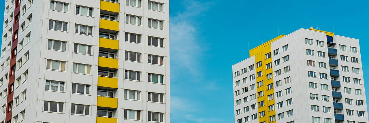 Zwei weiße Plattenbauten mit gelben und blauen Details. Beide Gebäude sind nur teilweise abgebildet, dazwischen ist blauer wolkenloser Himmel.