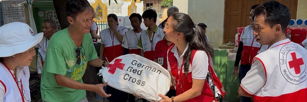Auf dem Bild sieht man eine Szene nach dem Erdbeben 2025 in Myanmar: Eine Mitarbeiterin des Deutschen Roten Kreuzes überreicht einen Sack voller Hilfsgüter an Mitarbeiterinnen und Mitarbeiter des Myanmarischen Roten Kreuzes. Auf dem Sack steht geschrieben "German Red Cross". Die Beteiligten schauen sich gegenseitig freudig an. Die Mitarbeiterinnen und Mitarbeiter des Myanmarischen Roten Kreuzes tragen Westen ihrer Hilfsorganisation, während die Mitarbeiterin des Deutschen Roten Kreuzes neutral gekleidet ist.