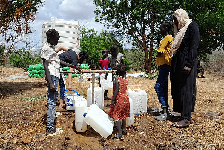 Mehrere Personen stehen mit Wasserkanistern um eine Wasserversorgungsstelle