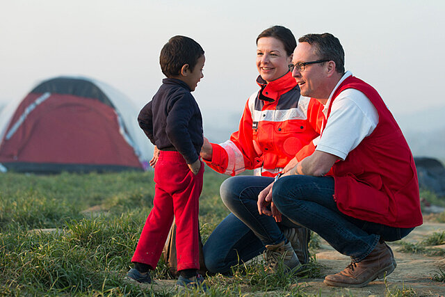 Foto: DRK-Mitarbeiter knien vor einem Fllüchtlingskind und sprechen mit ihm.