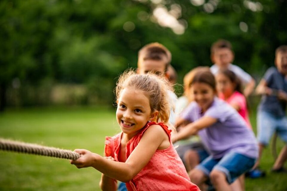 Kinder spielen Tauziehen, man sieht sechs Kinder auf einer Wiese an einem Seil ziehen. Das Mädchen ganz vorne stemmt sich lachend zurück, sie trägt ein rosa T-Shirt. Die Kinder hinter ihr sind nur verschwommen erkennbar.