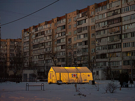 Beleuchtetes Wärmezelt des Ukrainischen Roten Kreuzes im Schnee und vor Hochhäusern ohne Strom