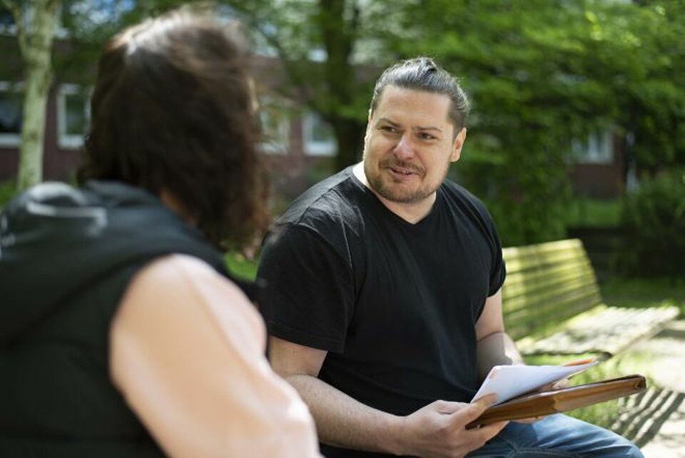 Ein Mann mit zusammengebundenem Haar und schwarzem T-Shirt sitzt im freien auf einer Bank. Er hält einige Dokumente in der Hand und spricht mit einer anderen Person. Sie sitzt neben ihm, mit dem Rücken zur Kamera. 