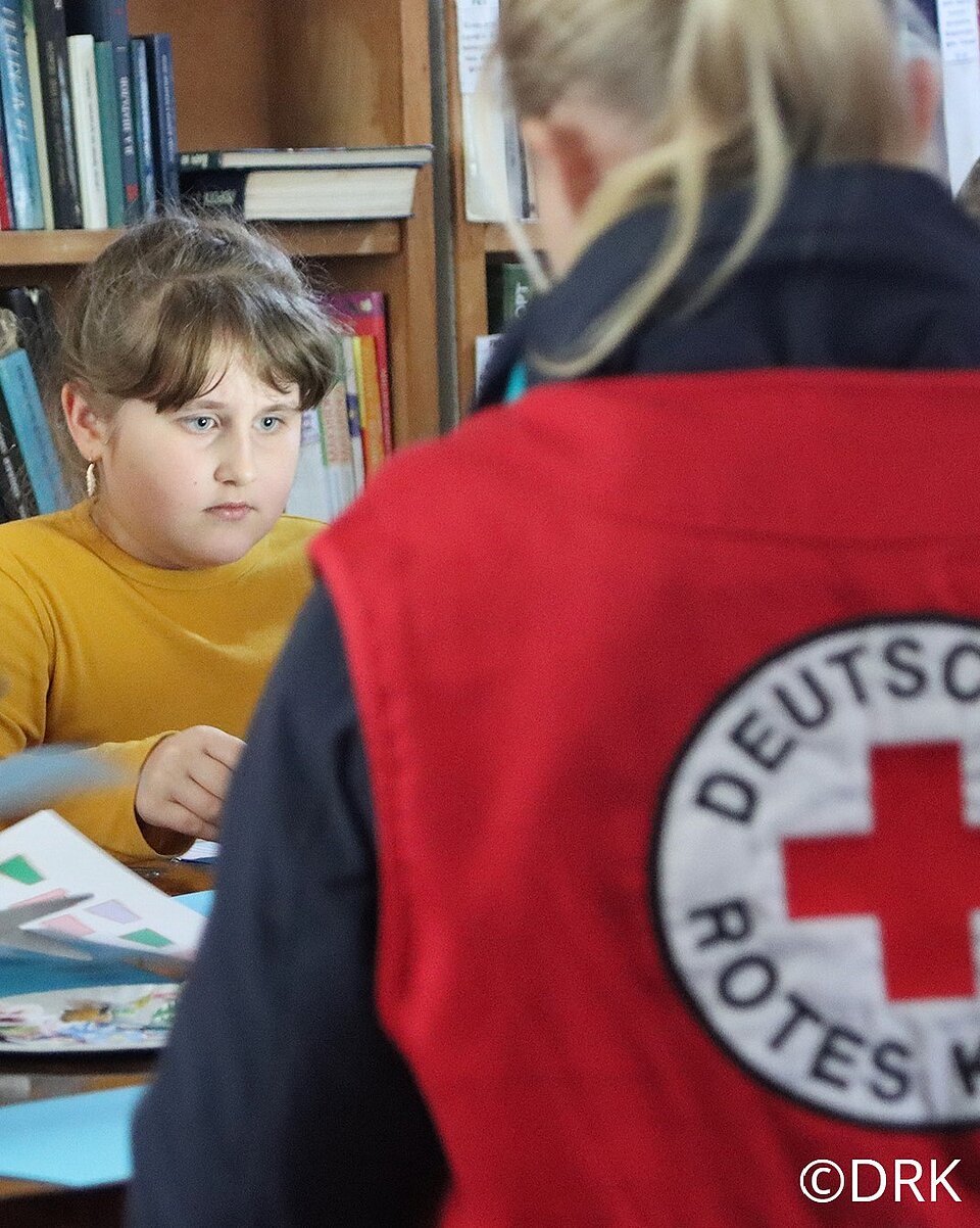 Im Vordergrund des Bildes sieht man eine Person von hinten. Sie trägt eine rote Weste mit der Aufschrift Deutsches Rotes Kreuz und blonde Haare zu einem Pferdeschwanz gebunden. Im Hintergrund sitzt ein Mädchen mit braunen Haaren und senfgelbem Pulli an einem Tisch. Es arbeitet konzentriert an einem Bild. Das Bild ist in der Ukraine entstanden.