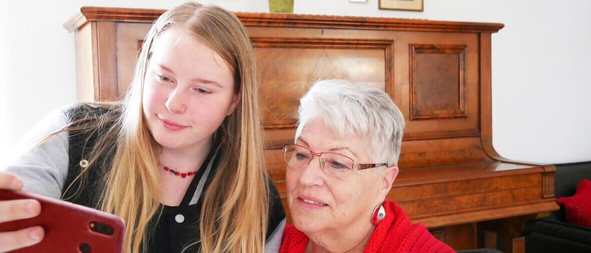 Eine Jugendliche und eine ältere Dame schauen in ein Smartphone, um ein Selfie zu schießen. Im Hintergrund steht ein Klavier.