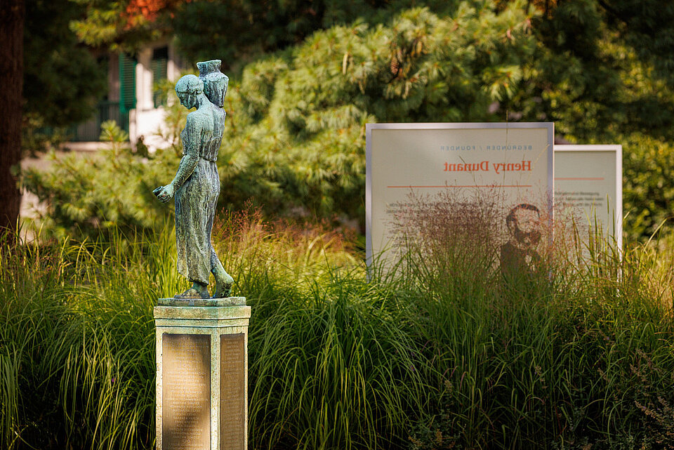 In einem Garten steht eine Skulptur und im Hintergrund ein Schild mit der Aufschrift "Henry Dunant"