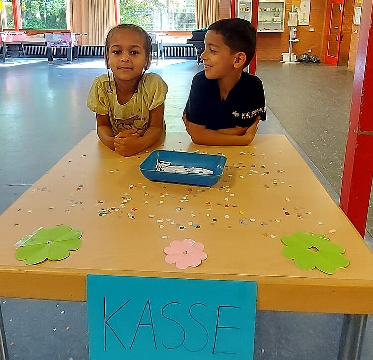 Ein Schüler und eine Schülerin der Vorschulklasse sitzen gemeinsam an einem Tisch, auf dem sie Lose verkaufen. Am Tisch hängt ein Danke-Schild. Der Vorschüler lächelt die Vorschülerin an, diese lächelt in die Kamera.