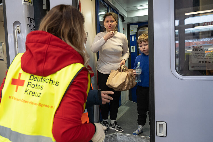 Eine Rotkreuzhelferin steht vor einem Zug und übergibt Tüten mit Lebensmitteln an Geflüchtete, die weiterfahren werden. In der Tür des Zugabteils stehen eine ukrainische Mutter und ihr Kind.