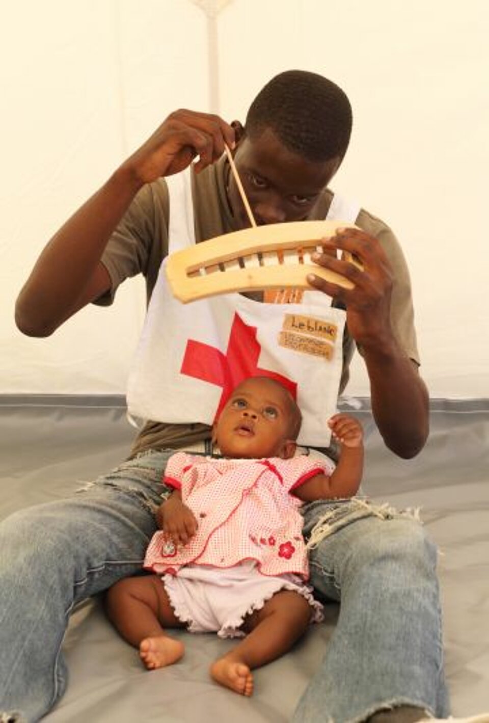 Foto: Sozialarbeiter Jerome sitzt mit einem Baby auf dem Schoß auf dem Boden und hält ihm ein Spielzeug hin