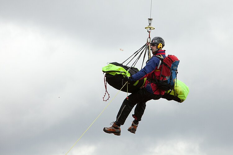 Eine Einsatzkraft hängt an einem Seil in der Luft und hält einen Verletzten in einer Trage. 