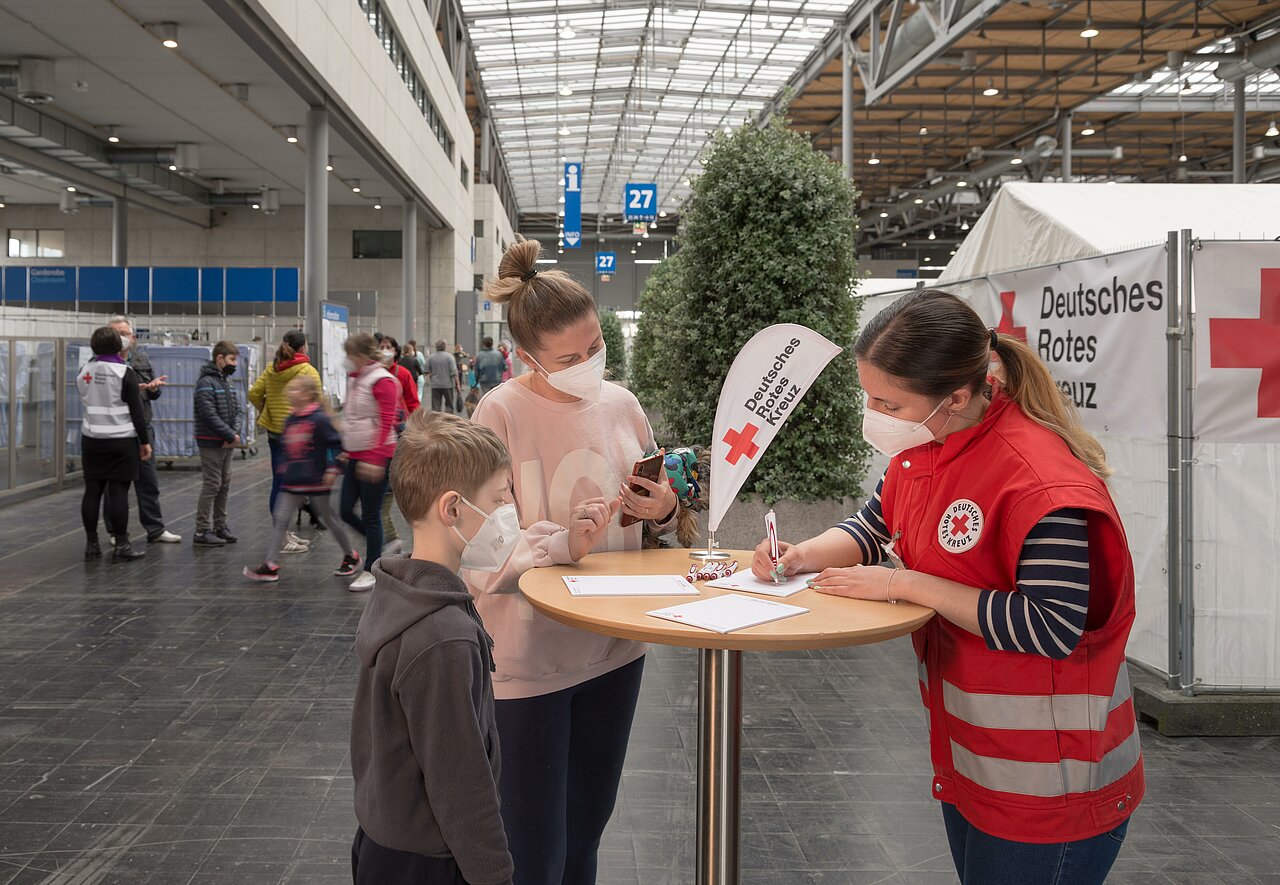 Ein Kind und eine Frau werden von einer DRK-Mitarbeiterin beraten. Sie stehen an einem Tisch in einer großen Halle.