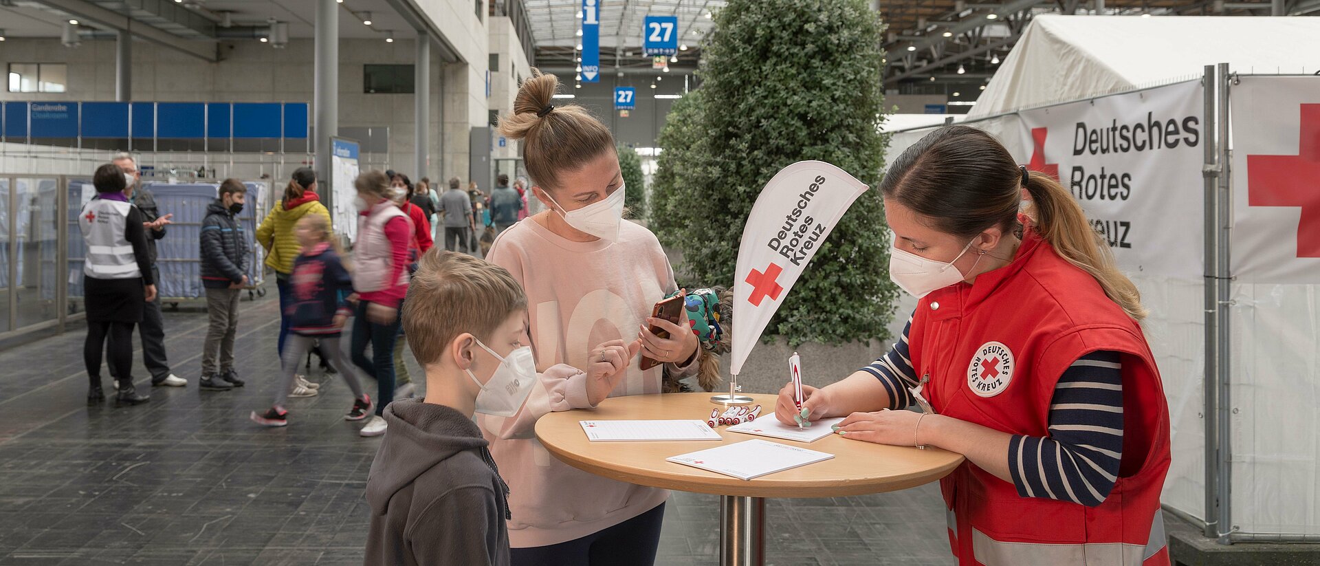 In einer großen halle stehen mehrere Menschen um einen Tisch. Eine Rotkreuzhelferin in roter Weste füllt ein Papier aus.