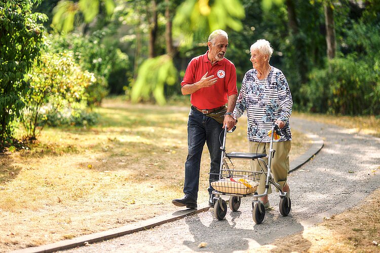 Pfleger und Seniorin mit Rollator spazieren durch Park