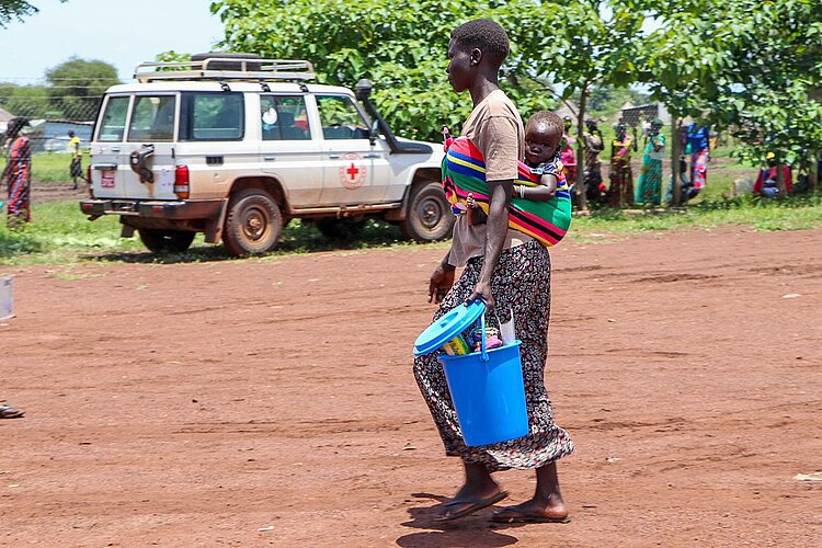 Geflüchtete Mutter und Baby mit Eimer vor Rotkreuz-Einsatzwagen in Uganda