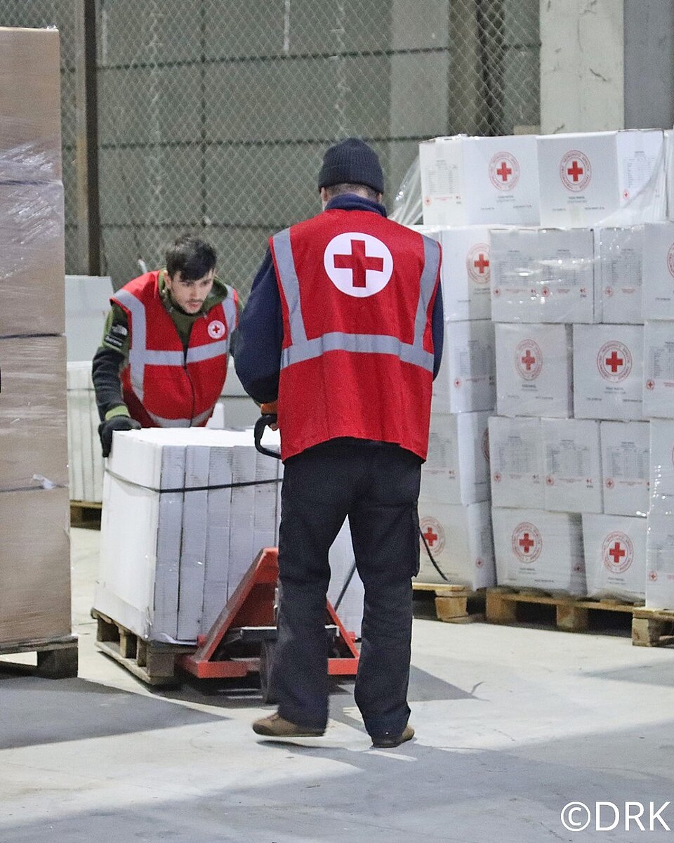 Zu sehen sind zwei Mitarbeiter vom Roten Kreuz der Ukraine. Sie befinden sich in einer Lagerhalle mit zahlreichen Kartons voller Hilfsgüter. Auf den Kartons ist das Rote Kreuz ebenso zu sehen, wie auf den Westen der beiden Mitarbeiter. Sie bewegen gerade eine Palette voller Kartons, einer schiebt und der andere zieht.