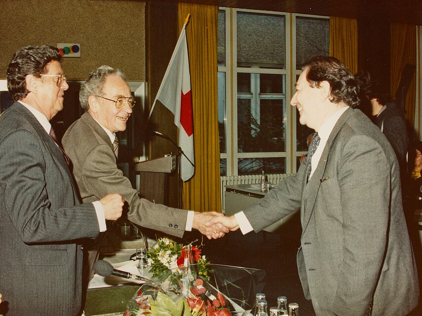 Drei Männer geben sich die Hand. Handschlag zwischen Prof. Dr. Christoph Brückner (re.), Präsident des DRK der DDR, und dem Vorsitzenden des Präsidialrats des westdeutschen DRK Dr. Josef Großmann. Li. der Präsident des DRK der Bundesrepublik Deutschland Botho Prinz zu Sayn-Wittgenstein