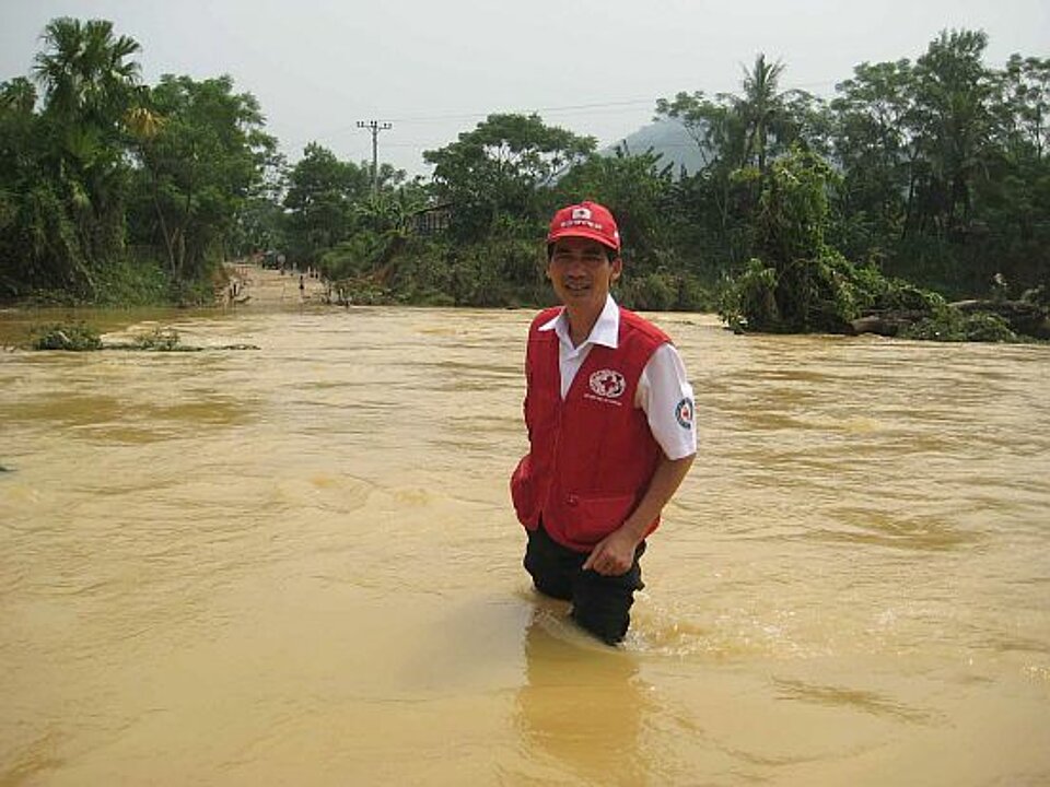 Foto: Ein asiatische aussehender Mann mit Rotkreuzmütze und Weste steht bis zur Hüfte in braunen Wassermassen