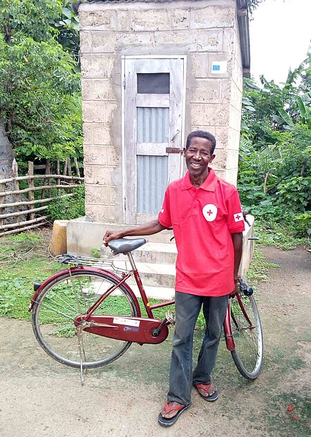 Rotkreuz-Freiwilliger mit Fahrrad vor Latrine