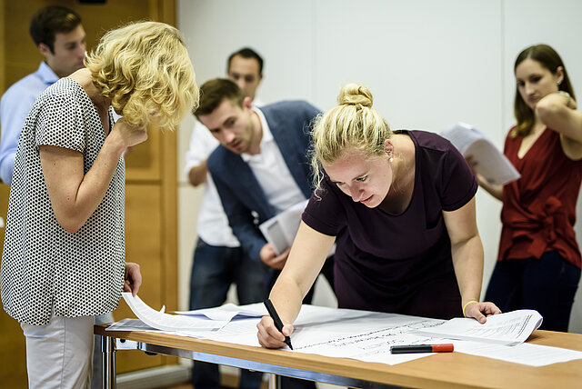 Eine blonde Frau schreibt auf ein Flipchart. Sie wird dabei von Teilnehmern eines Workshops beobachtet