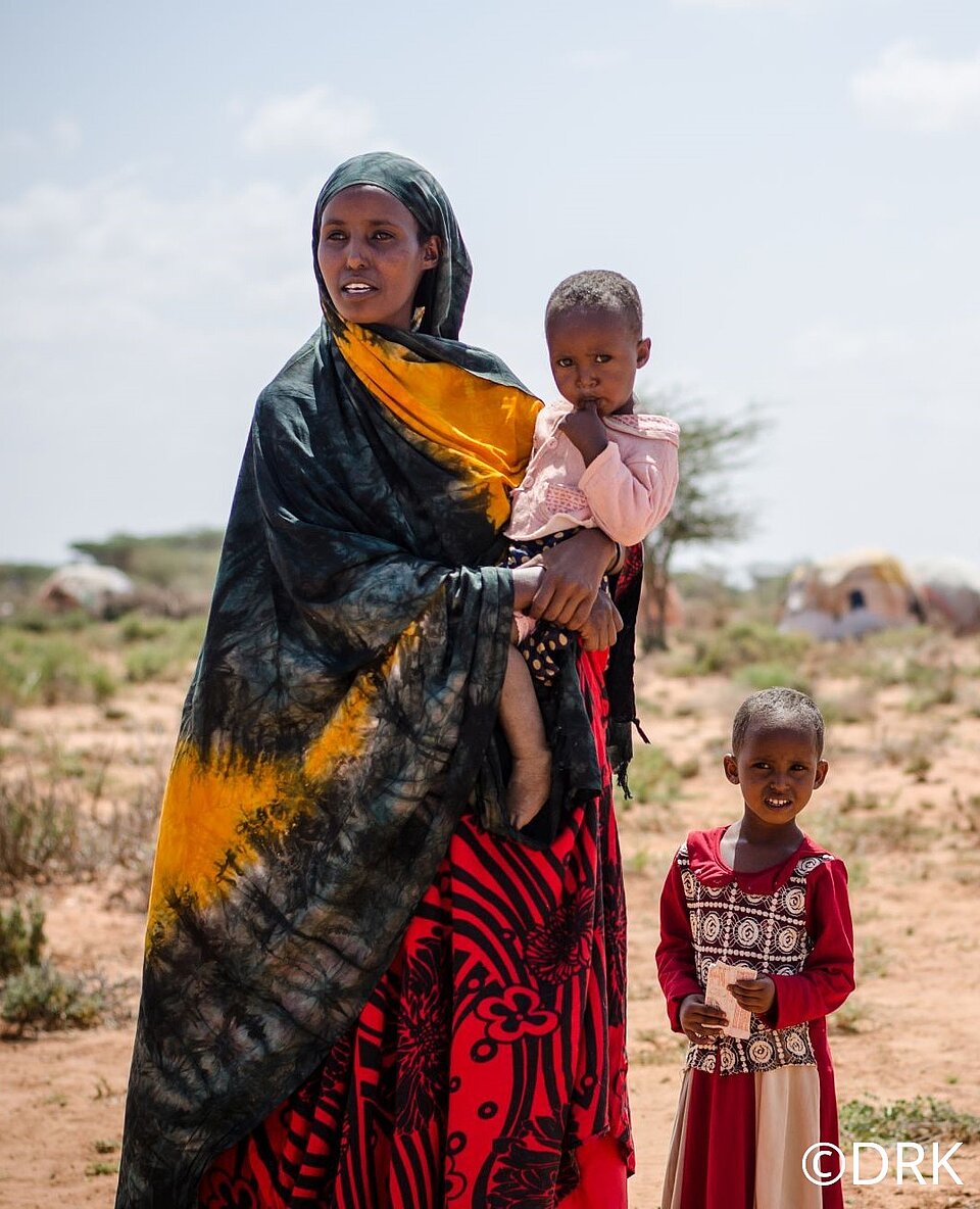 Das Bild ist in der Steppe von Somalia entstanden. Eine Frau und zwei Kinder in somalischer Kleidung schauen in die Kamera und in die Ferne. Die Frau trägt ein weites Kleid in den Farben rot, dunkelgrün und gelb. Eines der Kinder hält sie auf dem Arm. Es trägt eine rosane Bluse. Das andere Kind ist etwas älter, ca. 6-8 Jahre und trägt ein rot-weiß gestreiftes Kleid mit einem Muster. Rechts unten steht das Copyright-Symbol und der Text DRK.