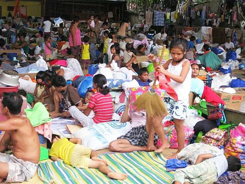 Foto: viele Kinder und vereinzelt auch Erwachsene sitzen dicht gedrängt auf dem Fußboden einer großflächigen Notunterkunft
