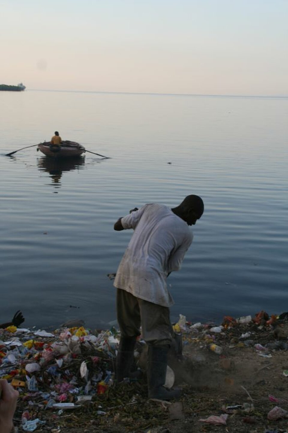 Foto: Ein Mann am Meeresufer im Sonnenuntergang, der im Mülle stochert; im Hintergrund ein Fischerboot auf dem Wasser