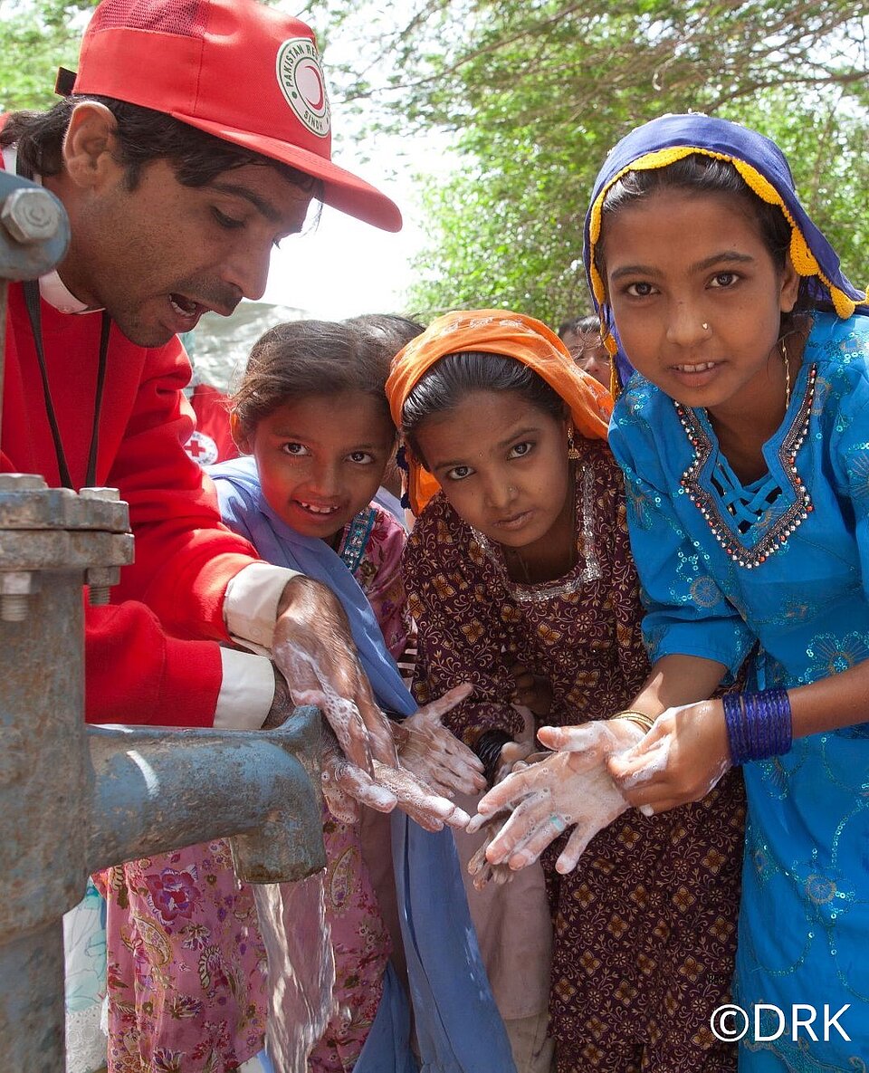 Auf dem Bild schauen drei Mädchen aus Pakistan in die Kamera. Sie tragen bunte Kleider und zwei von ihnen bunte Kopftücher. Neben ihnen steht ein Mann mit einer roten Jacke und roten Cap vom Pakistanischen Roten Halbmond. Er zeigt den Kindern an einem Brunnen, wie sie sich richtig die Hände waschen. Das Bild ist draußen und tagsüber entstanden. Rechts unten befindet sich ein Copyrightzeichen und das Kürzel DRK.
