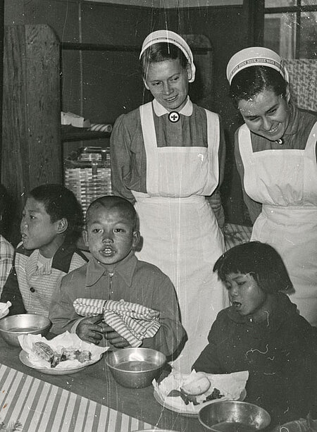 Foto: bedürftige koreanische Kinder beim Essen, hinter ihnen zwei deutsche Krankenschwestern