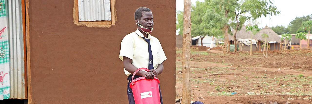 Eine ugandische Schülerin mit DRK-Eimer vor einem Haus