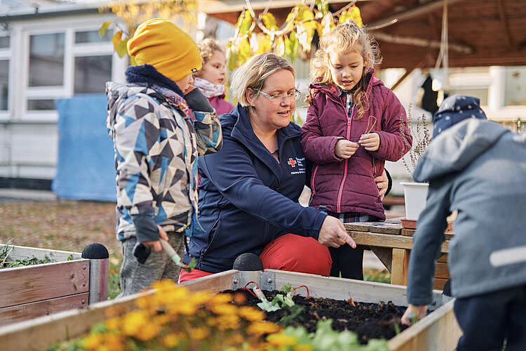 Nachhaltigkeit in DRK-Kitas Vier kleine Kinder stehen an einem Hochbeet und eine Erzieherin im blauen Shirt mit Rotkreuz-Logo zeigt auf etwas