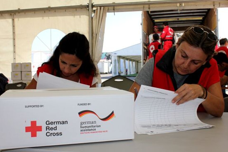 Zwei Mitarbeiterinnen des Hellenischen Roten KReuzes sitzen an einem Tisch und füllen Papiere aus. Vor ihnen leigt ein Hilfspaket mit dem Aufdruck "German Red Cross"