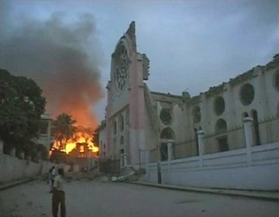 Foto eines großen Hauses, welches teilweise zerstört ist. Im Hintergrund sieht man die Flammen eines brennenden Nachbarhauses.