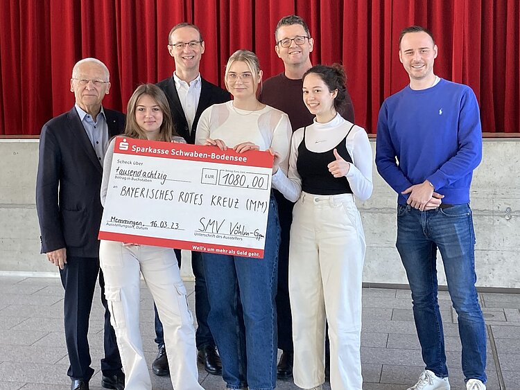 Auf dem Bild sind sieben Personen zu sehen. Bei ihnen handelt es sich um (von links nach rechts) hintere Reihe: Dr. Ivo Holzinger, Vorstand vom DRK Kreisverband Unterallgäu, Heinz Scheitzender vom KV Unterallgäu, Christian Herrmann (Schulleiter Vöhlin-Gymnasium) und Konstantin Heiß (Lehrer Vöhlin-Gymnasium). Alle vier Männer sind weiß und lächeln in die Kamera, drei von ihnen tragen Brillen, alle haben kurze Haare. Im Vordergrund stehen von links nach rechts die drei Schülersprecherinnen des Gymnasiums. Sie halten den sehr großen Scheck über 108 Euro in der Hand und lächeln in die Kamera.  