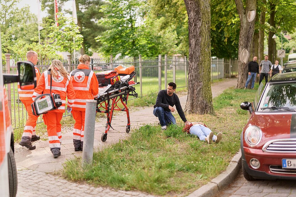 Erste Hilfe Situation nach einem Autounfall. Die Fahrerin wurde aus dem Auto geborgen. Der Rettungsdienst kommt nach Absetzen des Notrufs. Im Hintergrund Passanten