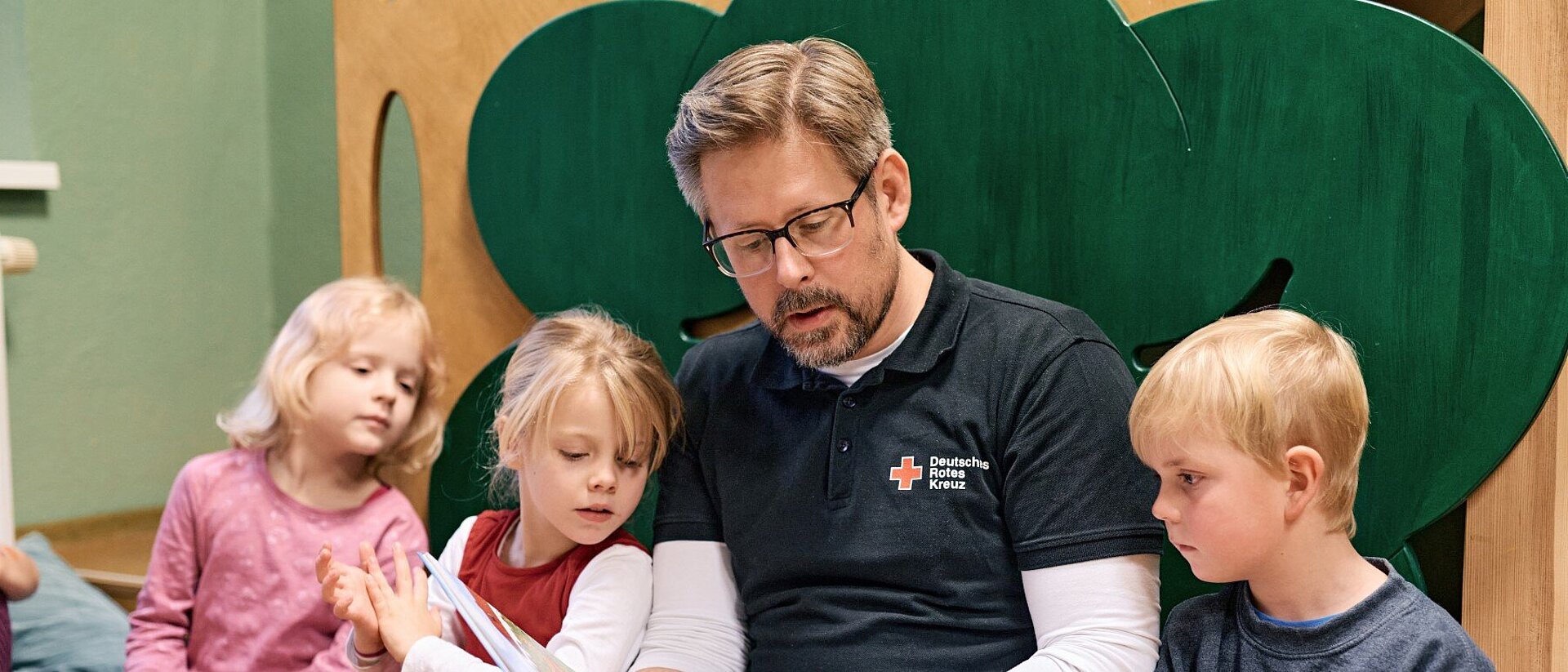 Ein Erzieher in einem DRK-Shirt liest drei Kindern, die neben ihm sitzen, etwas aus einem Buch vor.