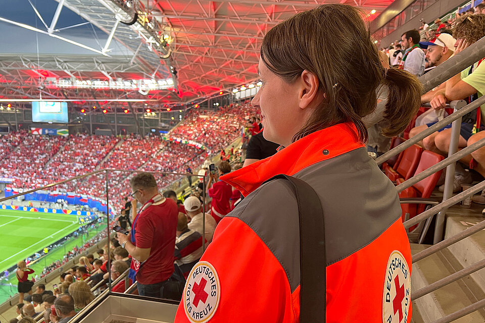 Eine Mitarbeiterin des Deutschen Roten Kreuzes in einer rot-grauen Jacke mit dem Logo des DRK auf dem Ärmel steht in einem Stadion und schaut auf das Spielfeld. Das Stadion ist voll besetzt mit Fans in rot-weißer Kleidung, die ein Fußballspiel verfolgen.